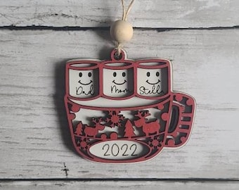 Może przedstawiać: Drewniana ozdoba świąteczna w kształcie kubka z trzema piankami. Pianki mają uśmiechnięte twarze i napisy "Dad", "Mom" i "Still". Kubek ma czerwono-biały wzór w kratę i rok "2022".