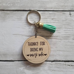 May include: Wooden keychain with a green tassel. The keychain has the text "Thanks for being my work wife" with two hearts.