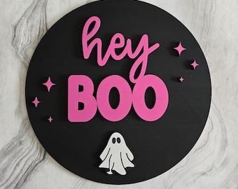 Può includere: Un cartello circolare in legno nero con le parole "hey BOO" in lettere rosa e un ritaglio di fantasma bianco. Il cartello è decorato con piccole stelle glitter rosa.