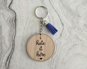 Può includere: Portachiavi in legno con un fiocco blu. Il portachiavi ha il testo "I hate it here" con un piccolo cuore.