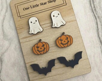 Può includere: Una scheda di esposizione in legno con il testo "One Little Star Shop" mostra un set di orecchini a tema Halloween. Gli orecchini sono a forma di fantasmi, zucche e pipistrelli. I fantasmi sono bianchi con occhi neri e sorrisi. Le zucche sono arancioni con occhi neri e un sorriso intagliato. I pipistrelli sono neri.