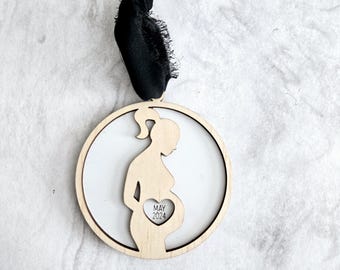 Può includere: Un ornamento in legno a forma di donna incinta con un cuore ritagliato nella pancia. Il cuore contiene il testo "MAY 2024". L'ornamento è attaccato a un nastro nero.