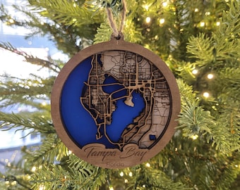 Può includere: Un ornamento natalizio in legno con una mappa blu e marrone di Tampa Bay, Florida. L'ornamento è appeso a un albero di Natale.