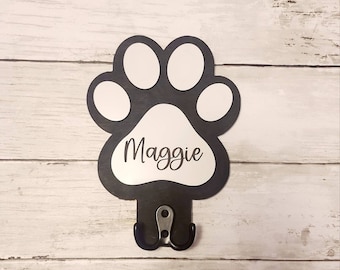 May include: Black paw print shaped hook with the name Maggie written in white on the front. The hook is mounted on a white wooden surface.