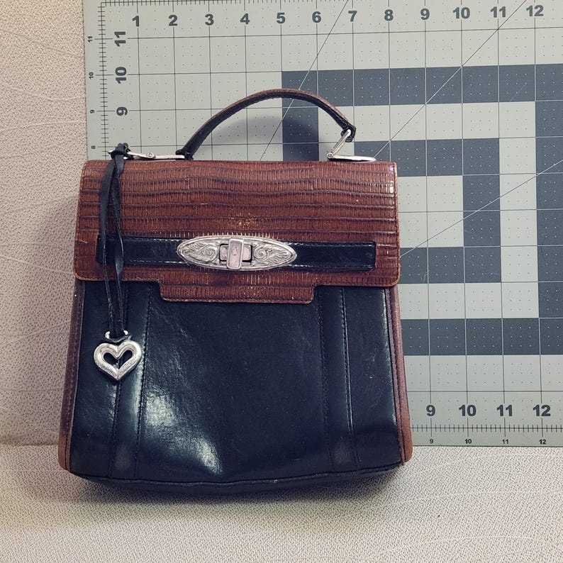 May include: A brown and black leather satchel handbag with a silver heart charm and a silver lock and key closure.
