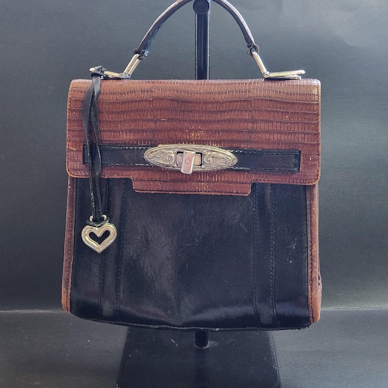 May include: A brown and black leather handbag with a silver heart charm and a silver clasp. The bag has a top handle and a shoulder strap.