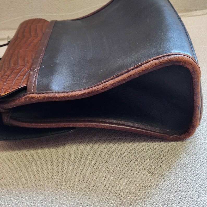 May include: A brown and black leather handbag with a flap closure. The bag has a brown leather trim and a brown leather strap.