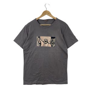 May include: A charcoal gray short-sleeve t-shirt featuring a rectangular graphic print on the chest. The print depicts a close-up illustration of a face wearing round, dark-rimmed glasses, focusing on the eyes and the bridge of the nose.