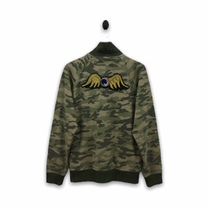 May include: A green, brown, and black camouflage patterned sweatshirt with a zipper. The sweatshirt has a large embroidered patch on the back with a yellow winged eye.