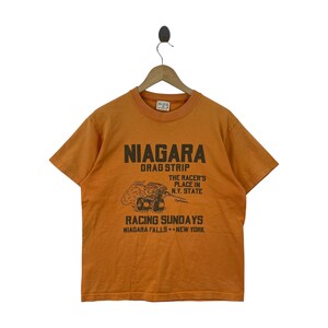 May include: An orange short-sleeve t-shirt featuring black text and a graphic of a drag racing car. The text reads NIAGARA DRAG STRIP, THE RACER'S PLACE IN N.Y. STATE, RACING SUNDAYS, and NIAGARA FALLS NEW YORK. The shirt is displayed on a wooden hanger.