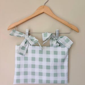 Può includere: Un top a quadretti bianchi e verde chiaro con spalline annodate, appeso a una gruccia di legno. La parte superiore presenta un motivo quadrato di quadrati verde chiaro su sfondo bianco. Le cinghie sono annodate in fiocchi.