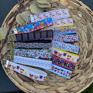 May include: A collection of colorful keychains with various Disney-themed designs. The keychains feature different characters, logos, and phrases, including "Never Too Old For Disney" and "Toy Story".
