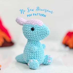 May include: A light blue crocheted dinosaur toy with pink accents. The toy is sitting on a white surface with a red and orange background. The text "No Sew Dinosaur PDF PATTERN" is written above the toy.