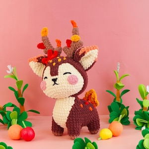 May include: A crocheted deer plush toy featuring a dark brown body with cream-coloured accents on the face and chest. The deer has pink cheeks, closed eyes, and antlers decorated with small orange and red yarn leaves. It stands upright.