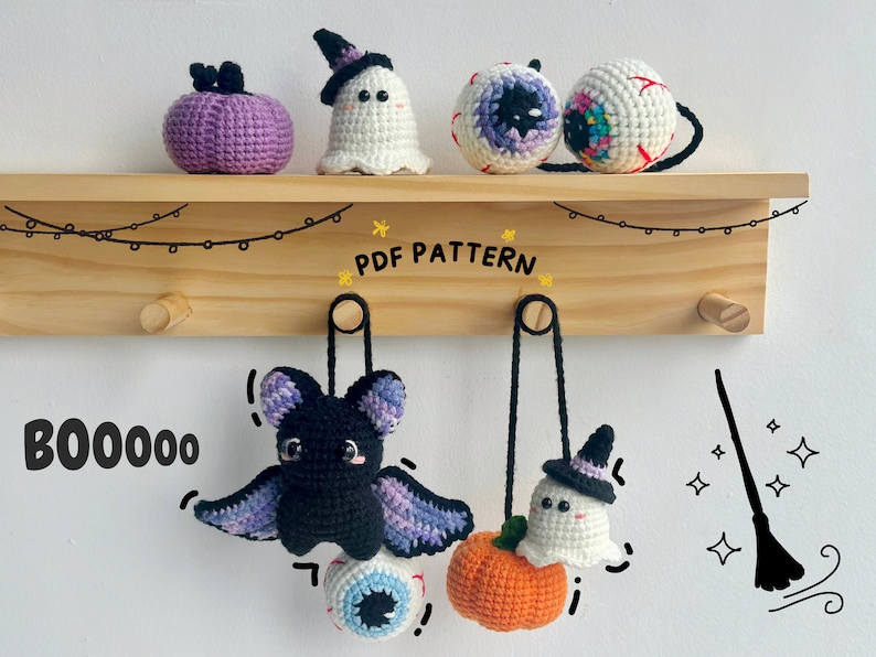 May include: A set of crocheted Halloween decorations hanging on a wooden rack. The decorations include a purple pumpkin, a white ghost, a black bat, a white ghost with a witch's hat, and two colorful eyeballs. The text "BOO0000" and "PDF PATTERN" are written on the rack.