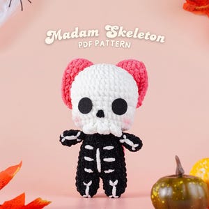 Pode incluir: Boneca de esqueleto de crochê com um crânio branco, corpo preto e detalhes rosa em forma de orelha. A boneca tem olhos pretos e um nariz preto. O texto "Madam Skeleton PDF Pattern" está acima da boneca. O fundo é rosa claro.