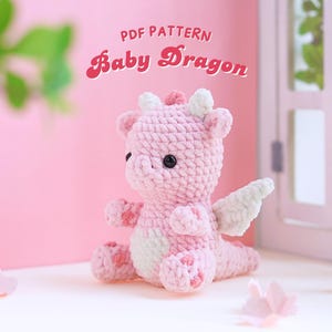 Może przedstawiać: Różowy, szydełkowy, mały smok z białymi skrzydłami. Smok ma czarne oczy i wzór w różowe i białe kropki. Tekst "PDF PATTERN Baby Dragon" znajduje się nad smokiem.
