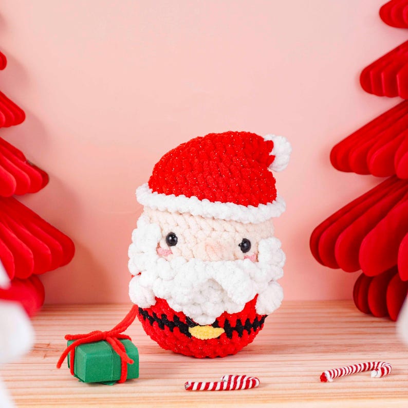May include: A crocheted Santa Claus figure with a red hat and suit, white beard and trim, and black belt with a yellow buckle. A small green gift box with a red ribbon is next to the Santa figure. Two candy canes are also present.