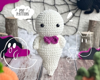 May include: A white crocheted ghost with a pink bow tie. The ghost has black eyes and a surprised expression. The ghost is sitting on a wooden surface with a pumpkin and a spiderweb in the background. The text "PDF PATTERN" is in a thought bubble above the ghost.