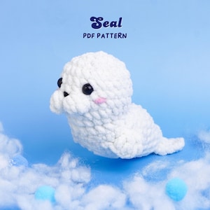 Könnte beinhalten: Ein weißes gehäkeltes Seehund-Plüschtier mit schwarzen Augen und rosa Wangen. Der Seehund befindet sich vor einem blauen Hintergrund mit dem Text "Seal PDF Pattern". Dekorative blaue Pompons und weiße Watte befinden sich im Vordergrund.