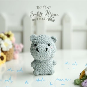 May include: A grey crocheted baby hippopotamus toy with black eyes and a pink nose. The toy is sitting on a white surface with a white picket fence in the background. The text "NO SEW Baby Hippo PDF PATTERN" is written in black cursive above the toy.