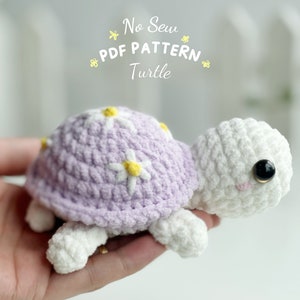 May include: A crocheted turtle amigurumi toy with a purple shell and white body. The shell has white daisies with yellow centers. The toy has black eyes and a pink mouth. The text "No Sew PDF PATTERN Turtle" is above the toy.