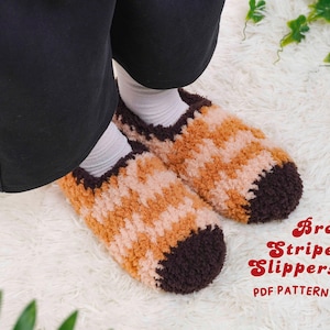 May include: A pair of brown and tan striped slippers with dark brown detailing. The slippers are made of a soft, textured material and are shown on a white, fluffy surface. Text on the image reads "Brown Striped Slippers PDF Pattern."