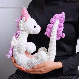 May include: A white crocheted dragon with pink accents. The dragon has a curled tail and a pink and white mane.