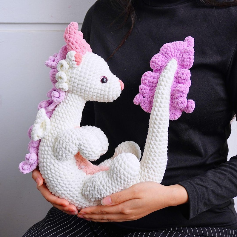 May include: A white crocheted dragon with pink accents. The dragon has a curled tail and a pink and white mane.