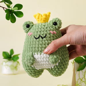 May include: A handmade green crochet frog toy with a yellow crown. The frog has black eyes, pink cheeks, and a white belly. The frog is a whimsical, soft toy.