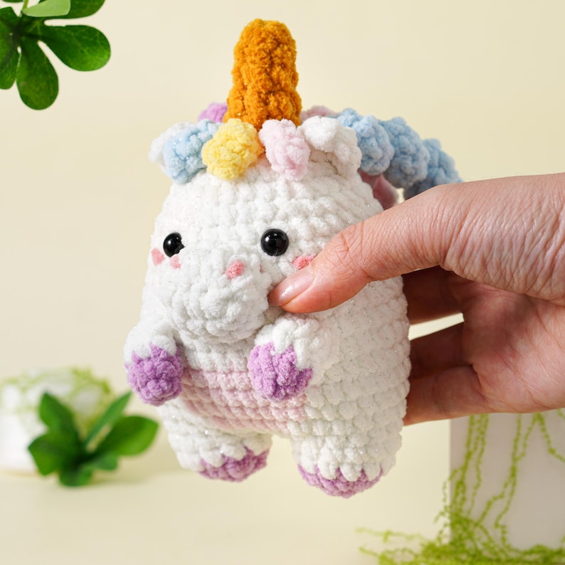 May include: A handmade white crochet unicorn with a yellow horn, pink cheeks, and multi-colored mane. The unicorn has black eyes and purple hooves. The plush toy is held by a hand.