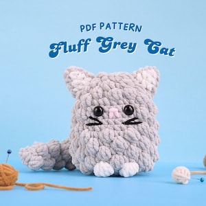 Puede incluir: Patrón PDF para un gato gris de crochet con patas blancas y una nariz rosa. El gato está sentado sobre una superficie azul con hilo y agujas.