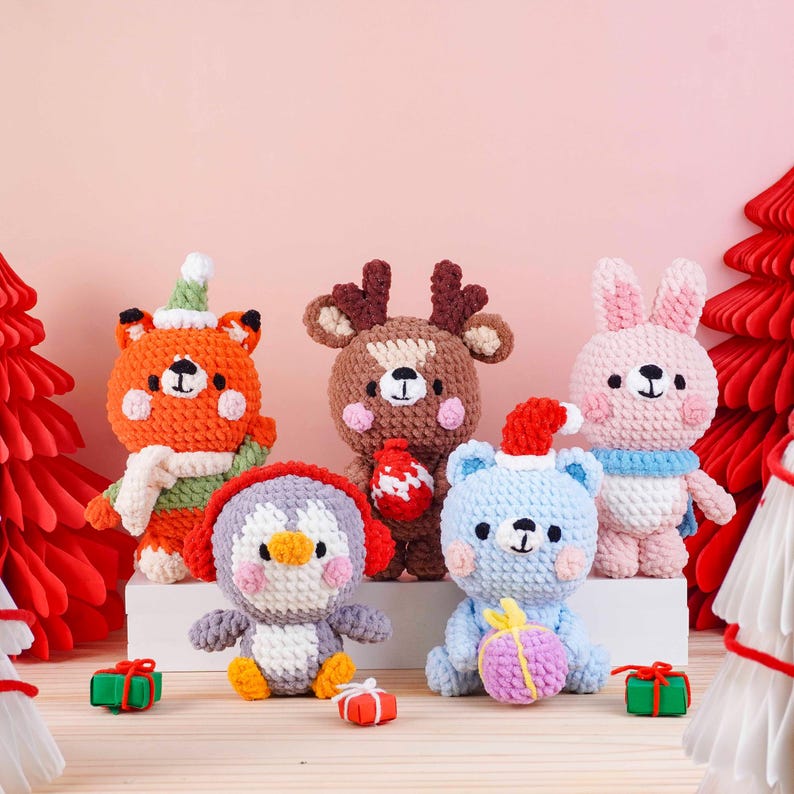 May include: A collection of five crocheted Christmas-themed plush toys. The toys include a fox, reindeer, penguin, bear, and bunny, each with unique festive details. The toys are arranged on a wooden surface with small gift boxes.