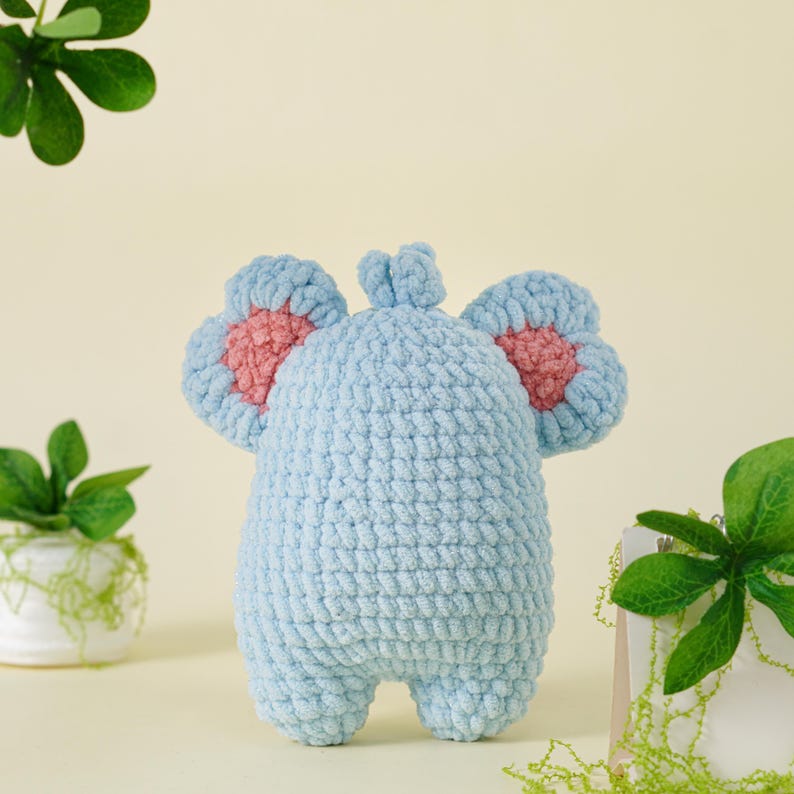 May include: A light blue crochet elephant toy with pink accents on the ears. The handmade stuffed animal is displayed against a light yellow background with green foliage.