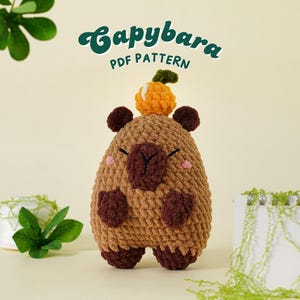 Op de afbeelding: Een gehaakte capybara knuffel in bruintinten, met donkerbruine accenten en een kleine oranje vrucht op zijn hoofd. De tekst "Capybara PDF Pattern" staat boven de knuffel.