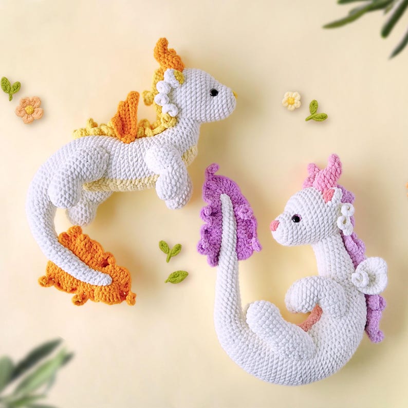 May include: Two crocheted dragon plushies, one with a yellow and orange tail and the other with a purple tail. Both dragons are white with pink accents.