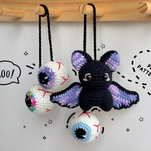 Pode incluir: Três decorações de Halloween de crochê penduradas em ganchos. Um morcego roxo e preto com um olho branco e um nariz rosa. Dois globos oculares brancos com detalhes coloridos. O texto "BOO!" está escrito em um balão de fala.
