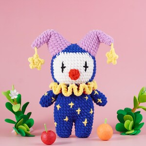 May include: A handmade blue and yellow crochet clown doll with a white face, red nose, and black cross eyes. The doll has a yellow ruffled collar, purple hat with yellow star accents, and yellow star details on the body. The background is pink.