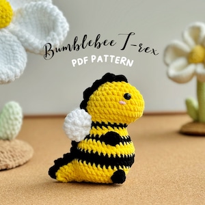 May include: A crocheted Bumblebee T-Rex amigurumi toy with yellow and black stripes, white wings, and black eyes. The toy is sitting on a brown surface with two crocheted white flowers in the background.