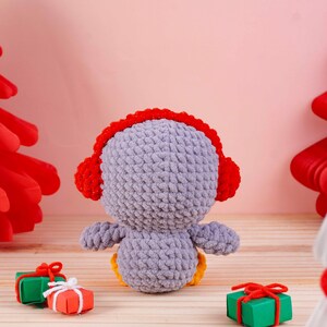 May include: A handmade, crocheted penguin doll in shades of gray, red, and yellow. The penguin wears red earmuffs and sits with its back to the viewer. Small gift boxes with red bows are in the foreground. The background is a soft pink.