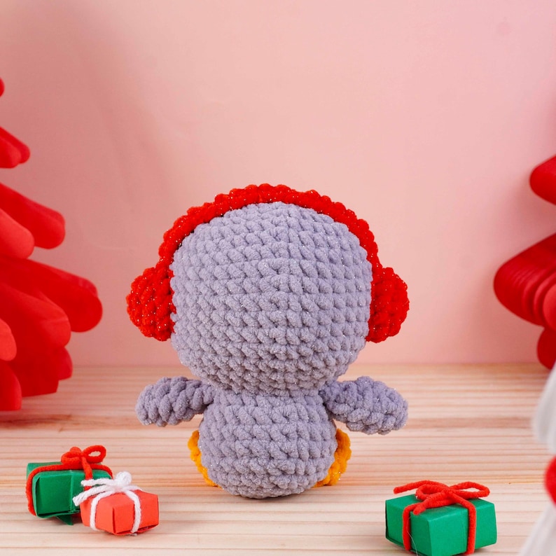 May include: A handmade, crocheted penguin doll in shades of gray, red, and yellow. The penguin wears red earmuffs and sits with its back to the viewer. Small gift boxes with red bows are in the foreground. The background is a soft pink.