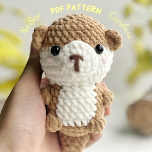 May include: A crocheted capybara plushie with brown and white yarn. The capybara has black eyes, pink cheeks, and a small brown nose. The text "PDF PATTERN Capybara" is in the background.