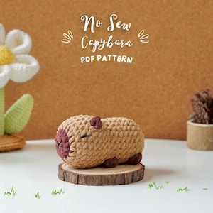 Puede incluir: Un capibara de crochet marrón y beige acostado sobre una rodaja de madera. El capibara tiene una nariz marrón y los ojos cerrados. El texto "No Sew Capybara PDF PATTERN" está sobre el capibara.
