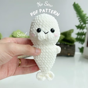 May include: White crocheted seal amigurumi toy with black eyes and pink cheeks. The toy is being held by a hand. The text "No Sew PDF Pattern" and "Seal" are visible in the image.