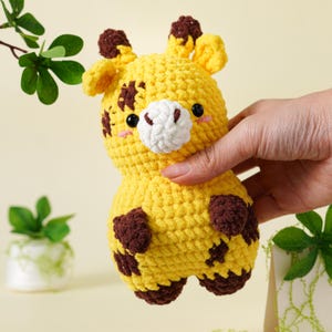 May include: A handmade yellow crochet giraffe toy with brown spots and accents. The giraffe has a white muzzle, black eyes, and small pink cheeks. The toy is held in a person's hand, showcasing its soft, textured appearance. The giraffe is a cute, cuddly toy.