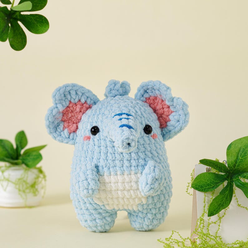 May include: A handmade, light blue crochet elephant with large ears featuring pink inner details. The elephant has a white belly, black eyes, and a trunk with blue lines. A cute, plush toy.