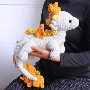 May include: A white crocheted dragon with yellow and orange crocheted accents. The dragon has a long tail and a small, curled up tail.