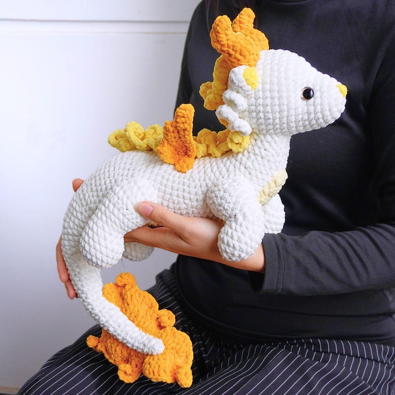 May include: A white crocheted dragon with yellow and orange crocheted accents. The dragon has a long tail and a small, curled up tail.