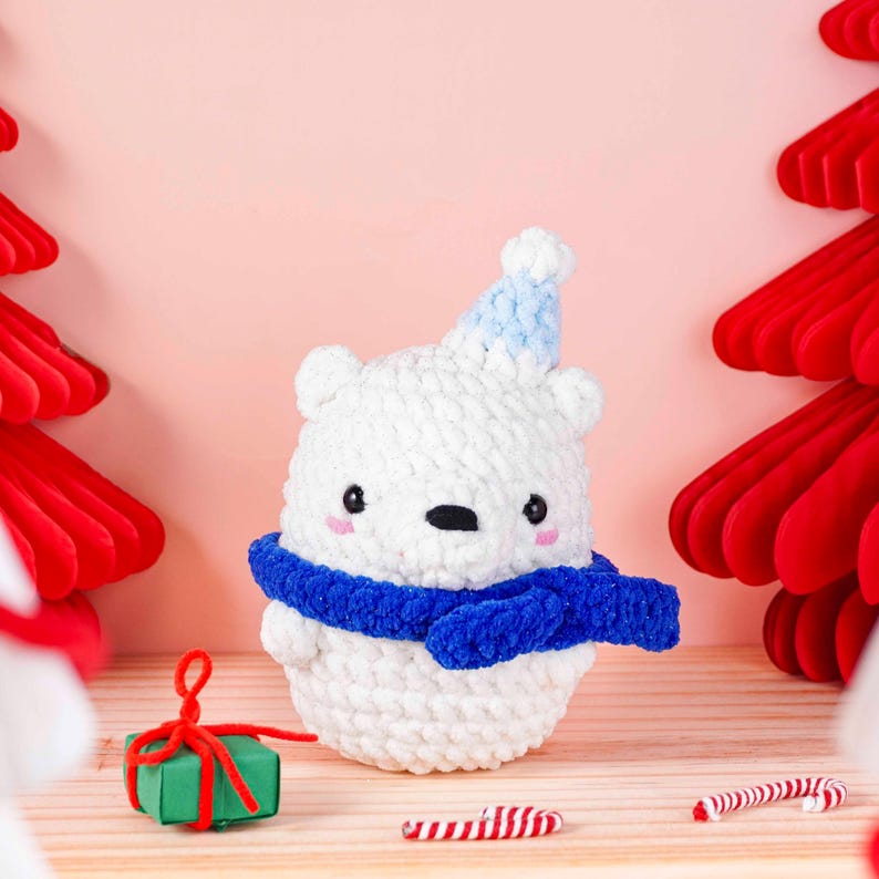 May include: A handmade white crochet polar bear wearing a blue scarf and a blue and white hat. The bear has black eyes and pink cheeks. A small green gift box with a red ribbon and candy canes are in the foreground. The background is a light pink.