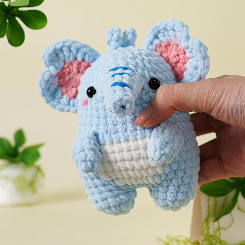 May include: A light blue crocheted elephant toy with pink inner ears and black eyes. The elephant has a white belly and a trunk. The toy is held by a hand.